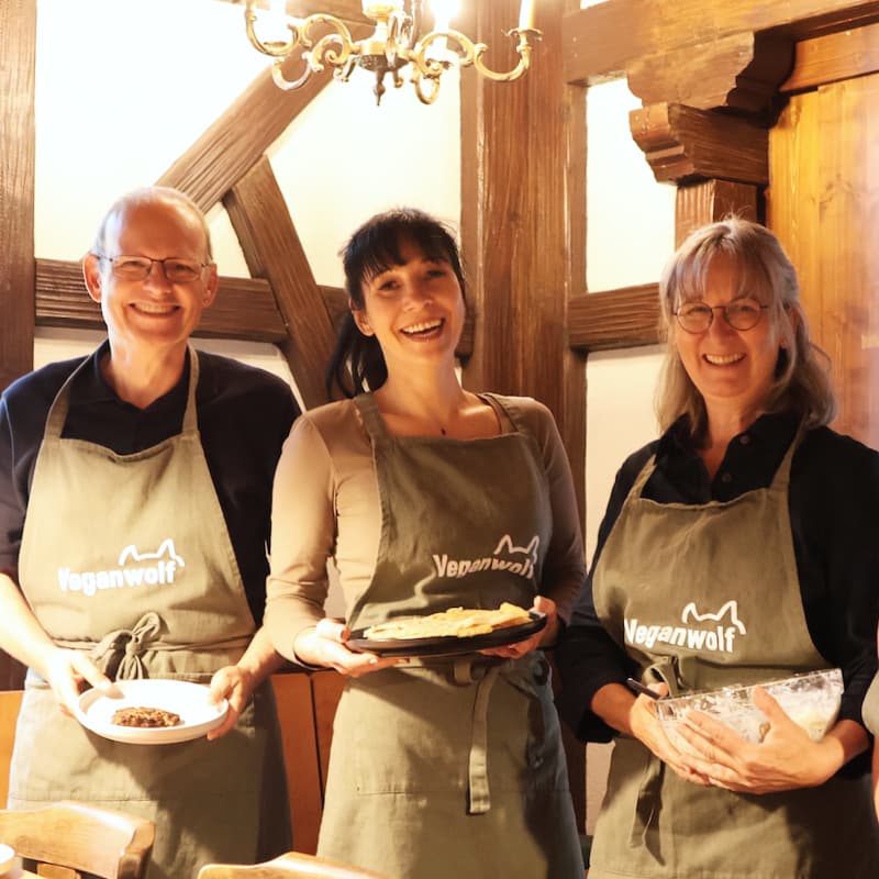 Drei Teilnehmende in Veganwolf-Schürzen zeigen ihre fertigen Gerichte vor der Fachwerkwand