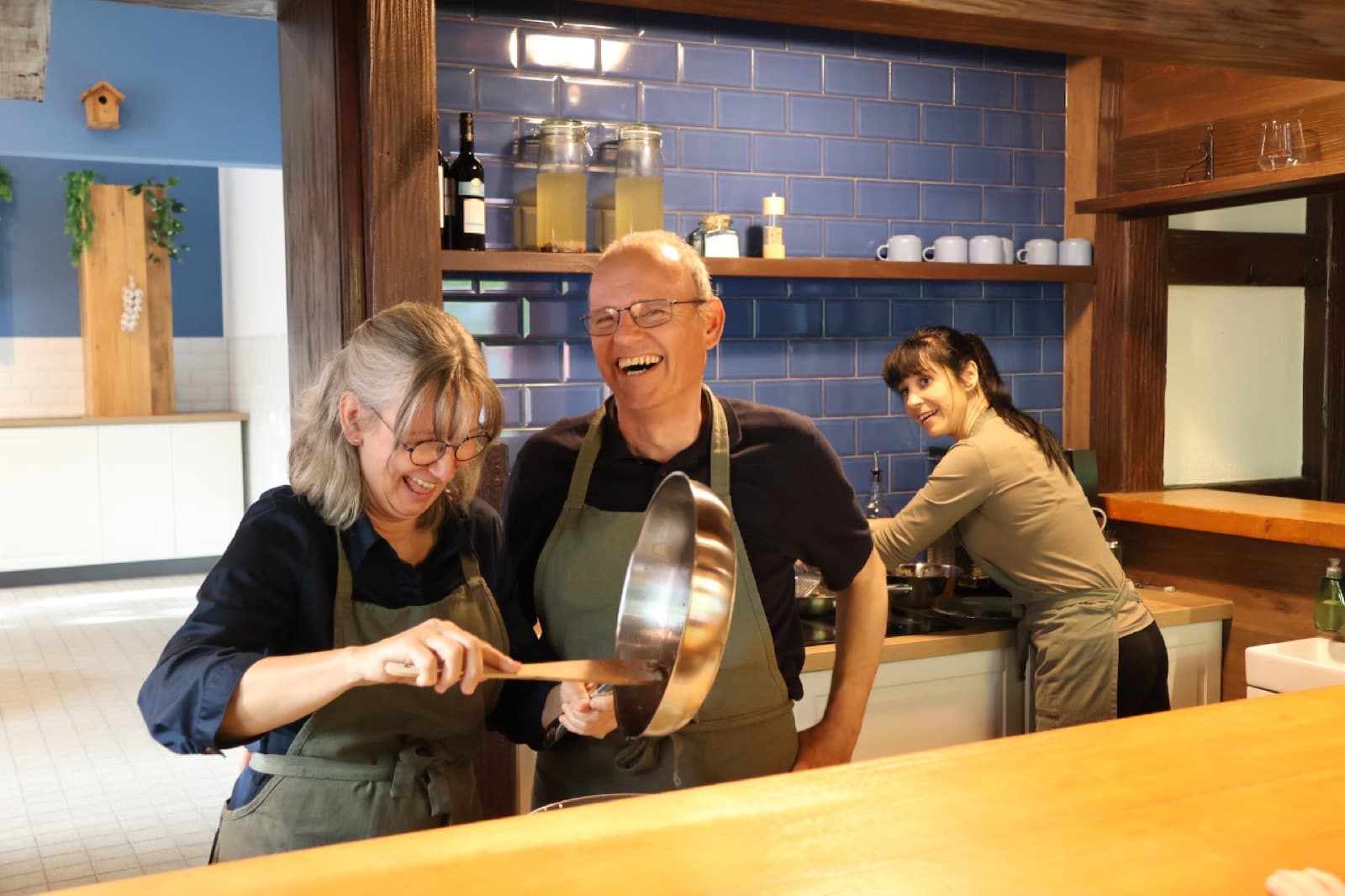 Zwei Teilnehmer lachen mit Pfanne und Pfannenwender an der Theke, im Hintergrund wird gekocht