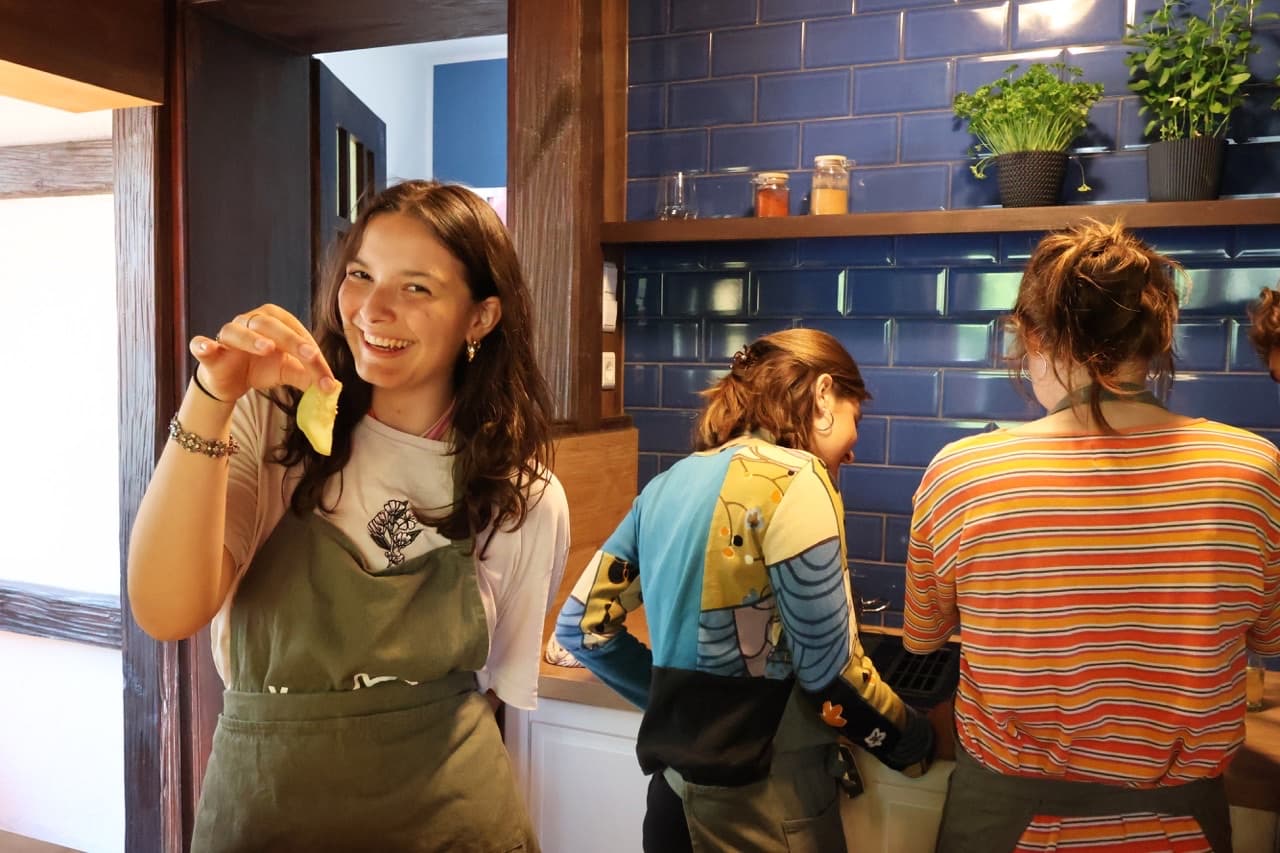 Drei Teilnehmende in Schürzen vor der blauen Fliesenwand, eine hält lachend frische Pasta hoch